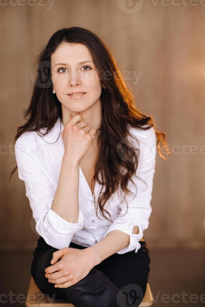 portrait de une content Jeune brunette femme dans une blanc chemise séance sur une chaise photo