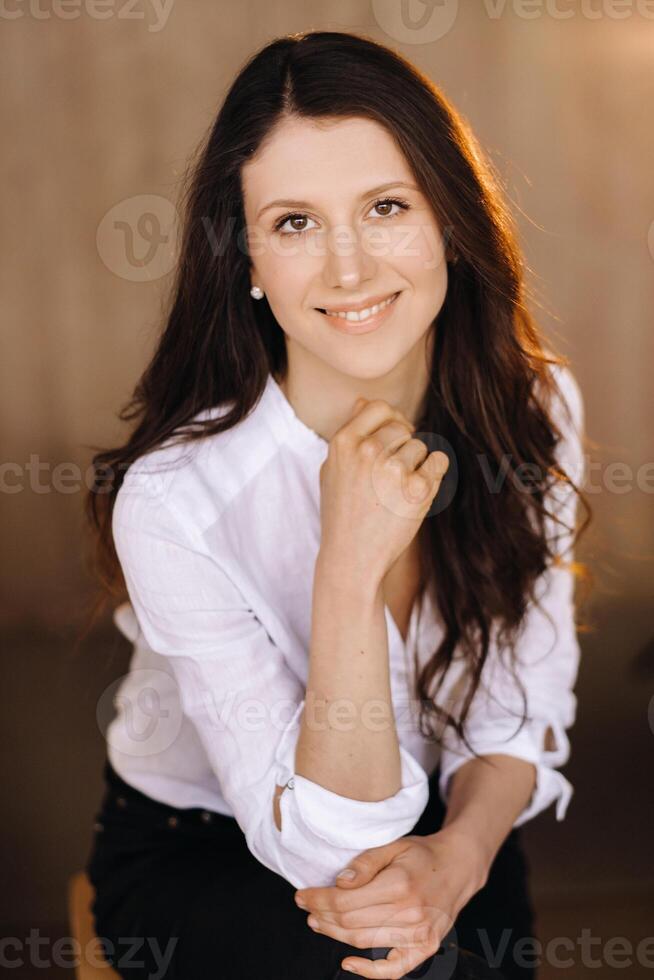 portrait de une content Jeune brunette femme dans une blanc chemise séance sur une chaise photo