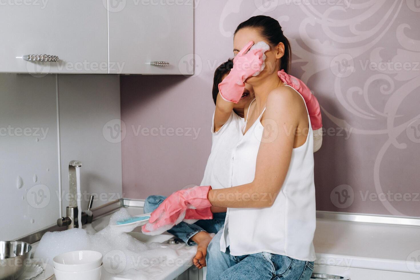 femme au foyer maman dans rose gants lavages vaisselle avec sa fils par main dans le évier avec détergent. une fille dans blanc et une enfant avec une jeter nettoie le maison et lavages vaisselle dans fait maison rose gants. photo