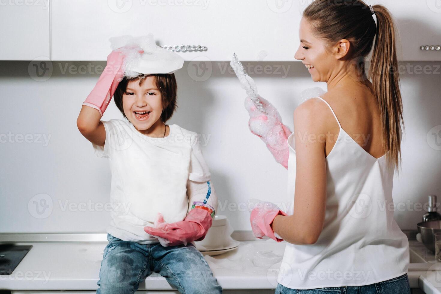 femme au foyer maman dans rose gants lavages vaisselle avec sa fils par main dans le évier avec détergent. une fille dans blanc et une enfant avec une jeter nettoie le maison et lavages vaisselle dans fait maison rose gants. photo