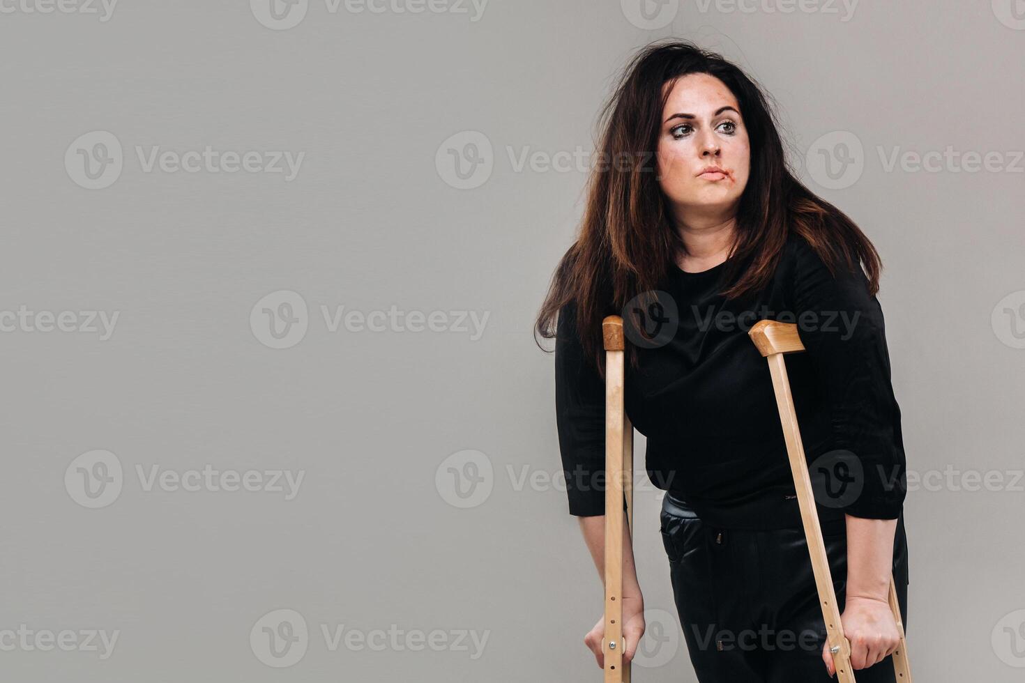 une femme battu dans noir vêtements avec béquilles dans sa mains sur une gris Contexte photo