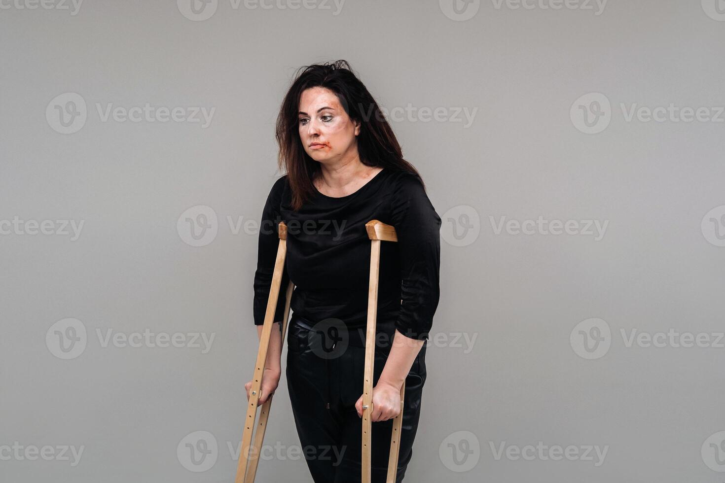 une femme battu dans noir vêtements avec béquilles dans sa mains sur une gris Contexte photo