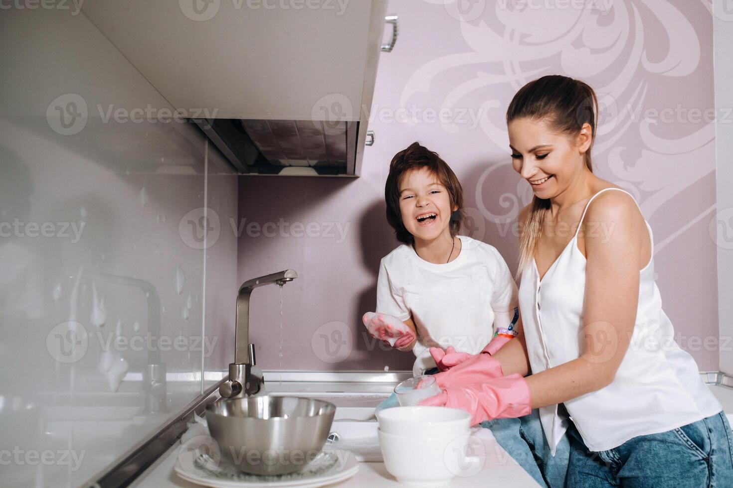 femme au foyer maman dans rose gants lavages vaisselle avec sa fils par main dans le évier avec détergent. une fille dans blanc et une enfant avec une jeter nettoie le maison et lavages vaisselle dans fait maison rose gants. photo