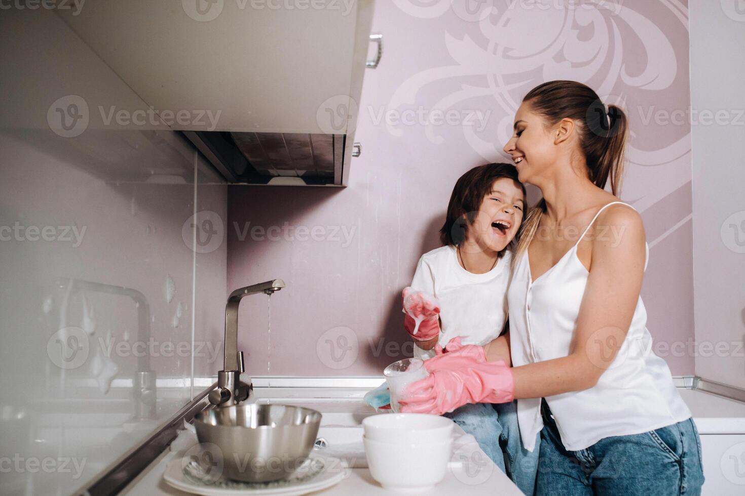 femme au foyer maman dans rose gants lavages vaisselle avec sa fils par main dans le évier avec détergent. une fille dans blanc et une enfant avec une jeter nettoie le maison et lavages vaisselle dans fait maison rose gants. photo