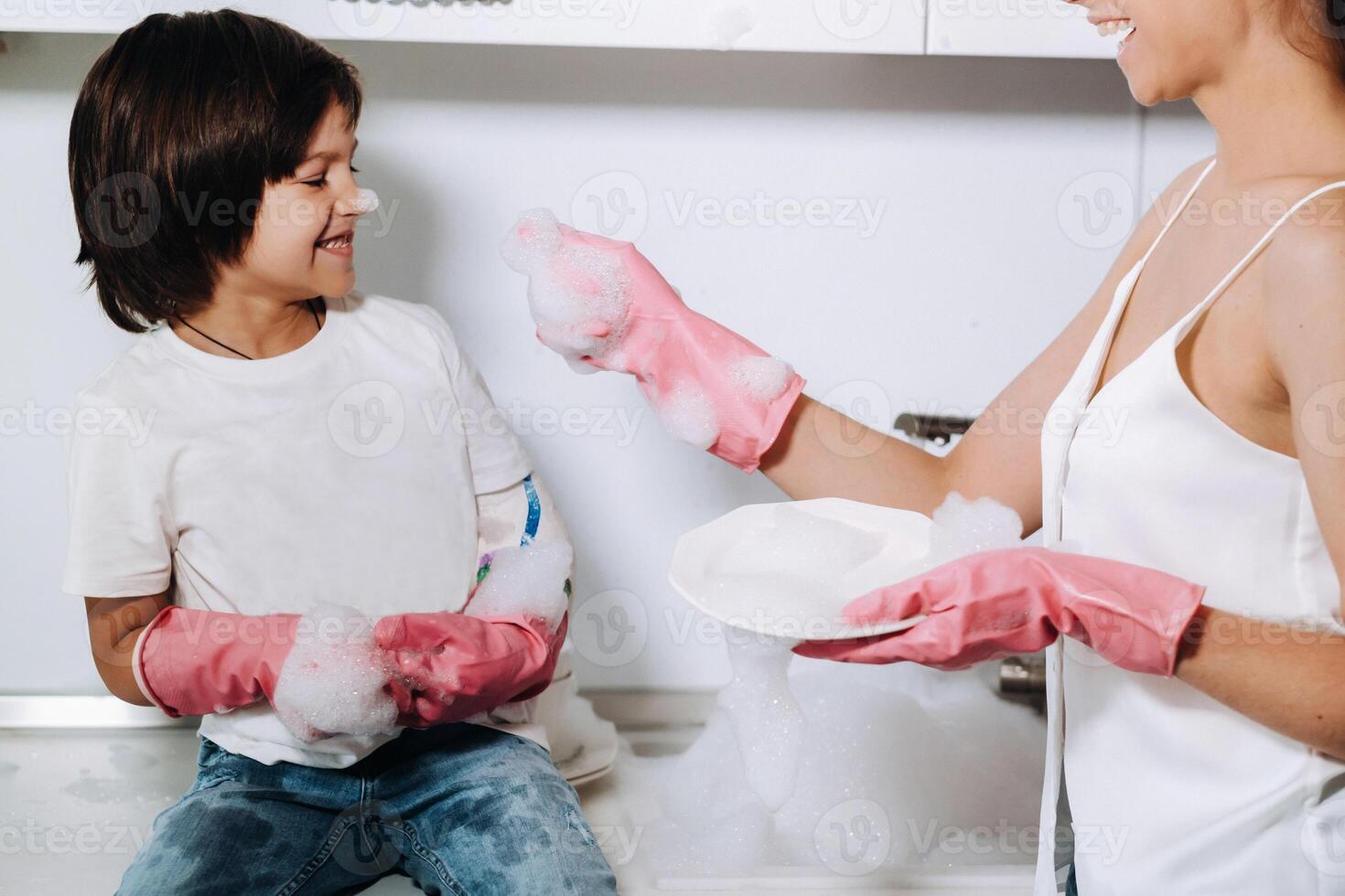 femme au foyer maman dans rose gants lavages vaisselle avec sa fils par main dans le évier avec détergent. une fille dans blanc et une enfant avec une jeter nettoie le maison et lavages vaisselle dans fait maison rose gants. photo