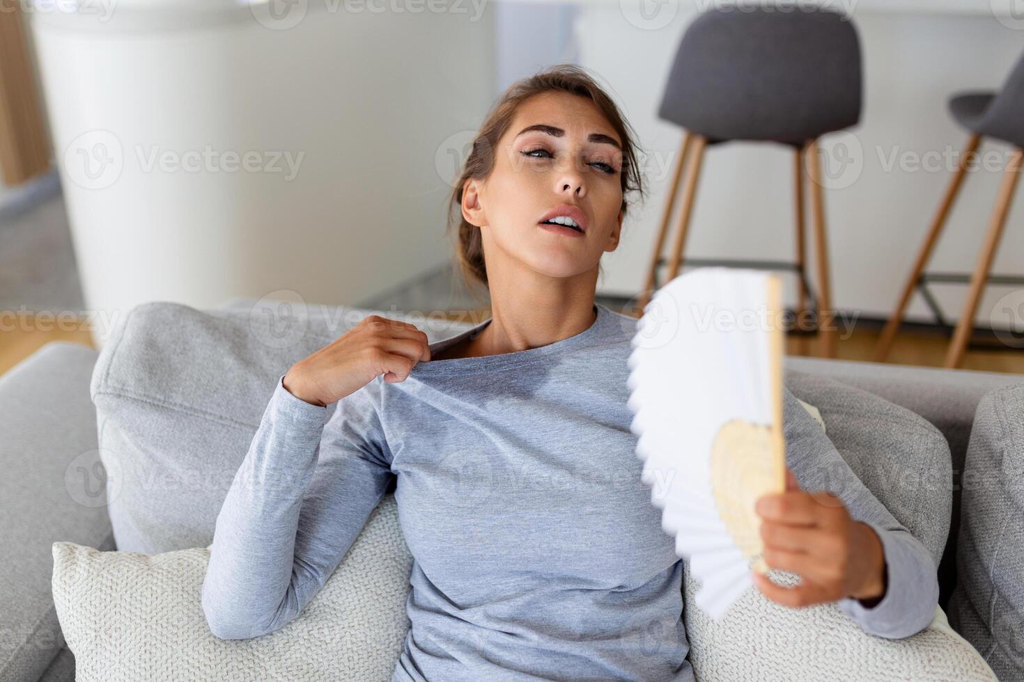 surchauffé femme asseoir sur canapé à Accueil ressentir chaud agitant avec main ventilateur refroidissement bas, transpiration fille se détendre sur canapé dans vivant pièce tenir vaciller souffrir de chaleur, non air Conditionneur système photo