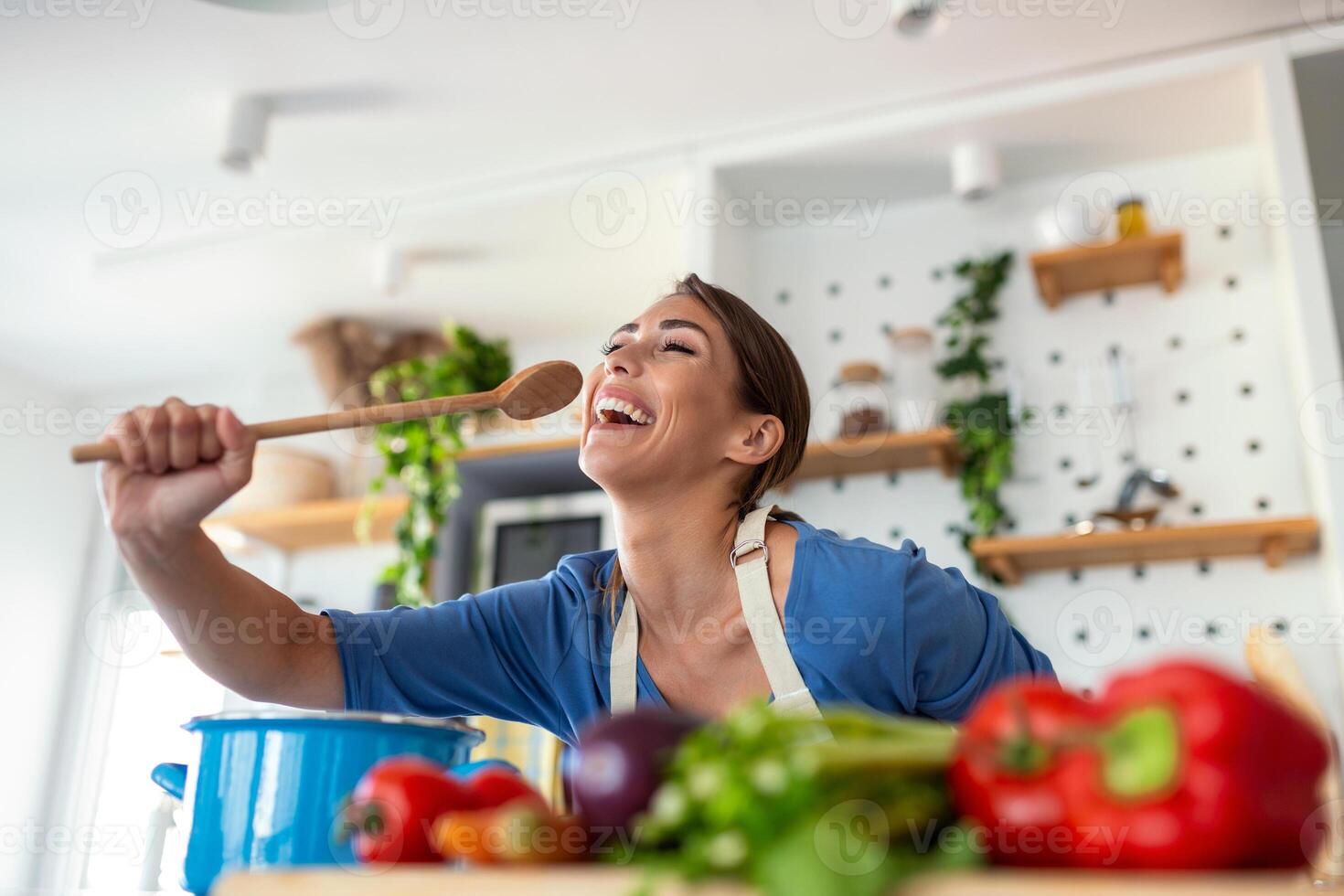 marrant magnifique femme en chantant dans spatule, cuisine dans moderne cuisine, en portant spatule comme microphone, dansant, écoute à musique, espiègle fille ayant amusement avec ustensiles de cuisine, en train de préparer aliments. photo