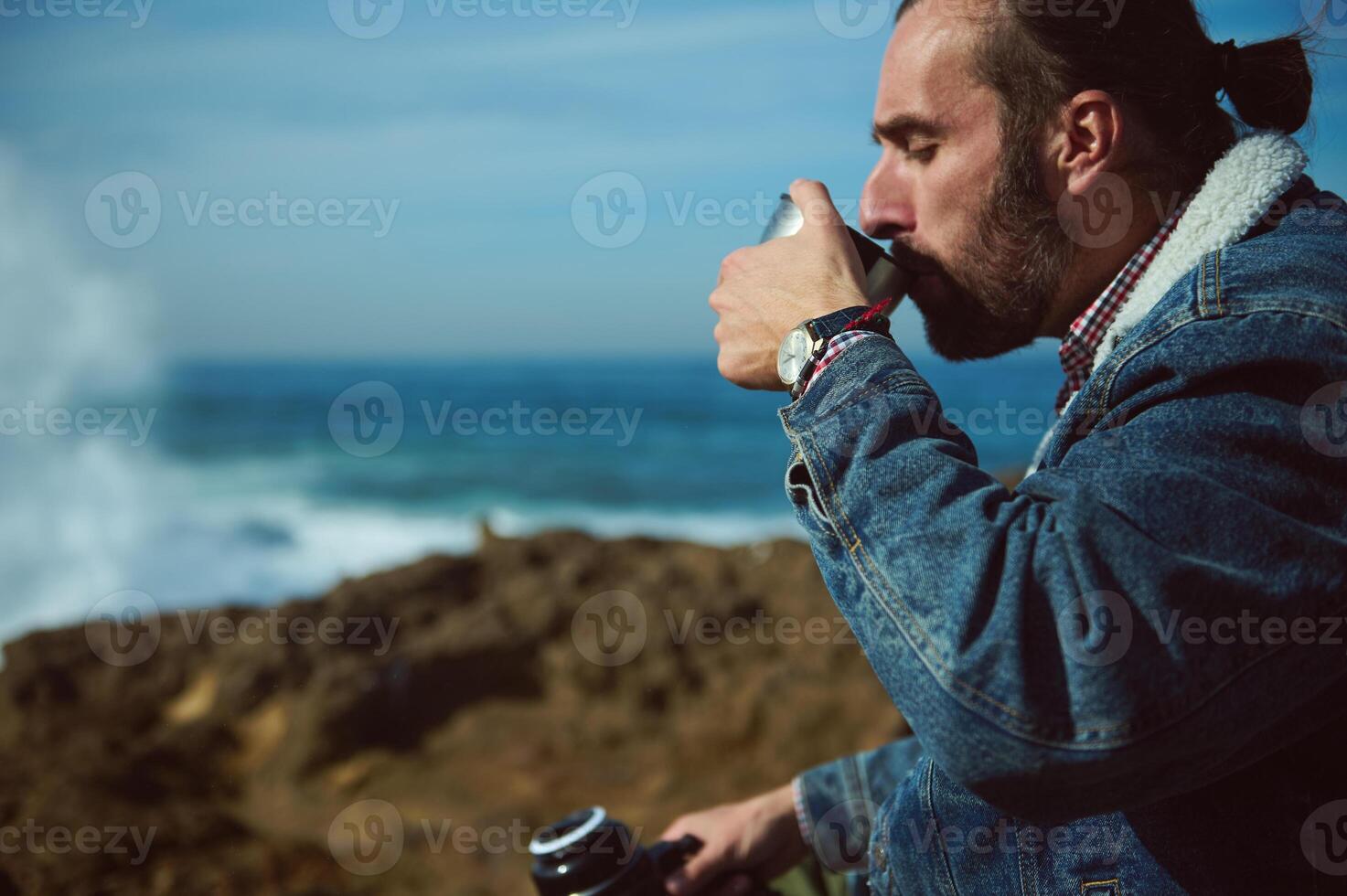 détendu barbu Masculin touristique voyageur aventurier séance sur une rocheux falaise par océan et en buvant l'eau de une thermos photo