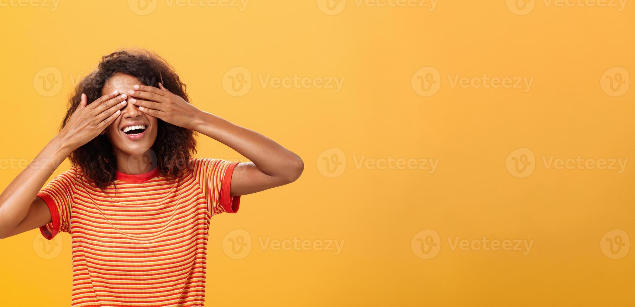 fermeture mon yeux et compte dix. portrait de charmant rêveur et content marrant africain américain aux cheveux bouclés femelle dans rayé branché T-shirt couvrant vue avec paume et souriant attendre surprise photo