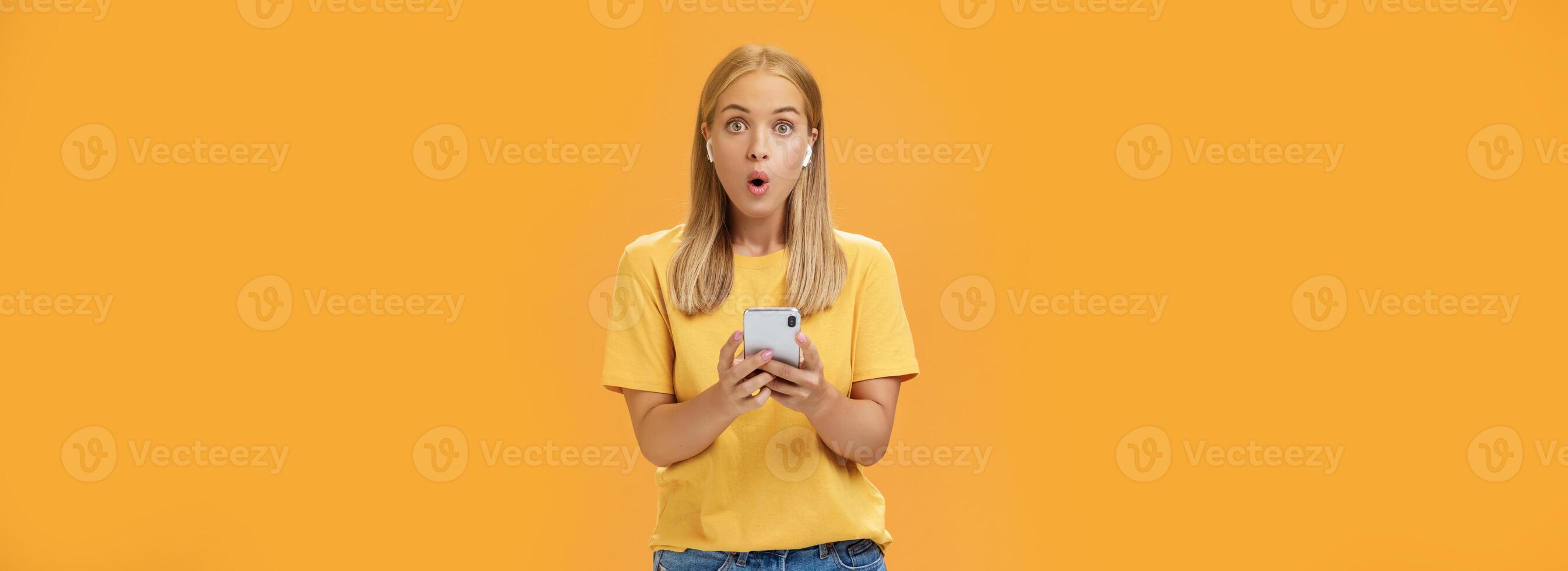 femme surpris Comment impressionnant des sons Nouveau sans fil écouteurs en portant téléphone intelligent portant écouteurs pliant lèvres dans sensationnel et stupéfaction écoute la musique impressionné par bien produit plus de Orange Contexte photo