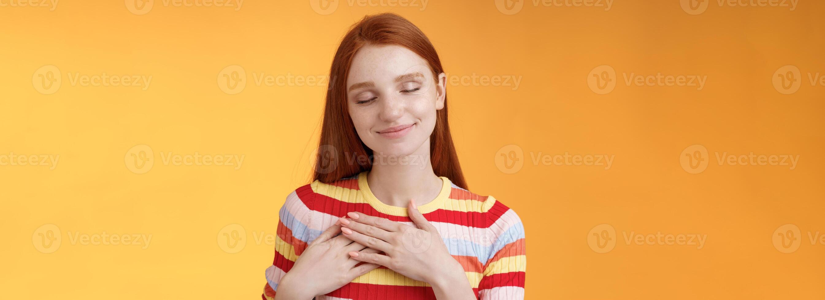 cœur garde chaud enregistrer très cher souvenirs à l'intérieur. souriant soumissionner réconfortant roux charmant fille toucher poitrine sourire fermé yeux rappeler charmant agréable moment permanent romantique Orange Contexte photo