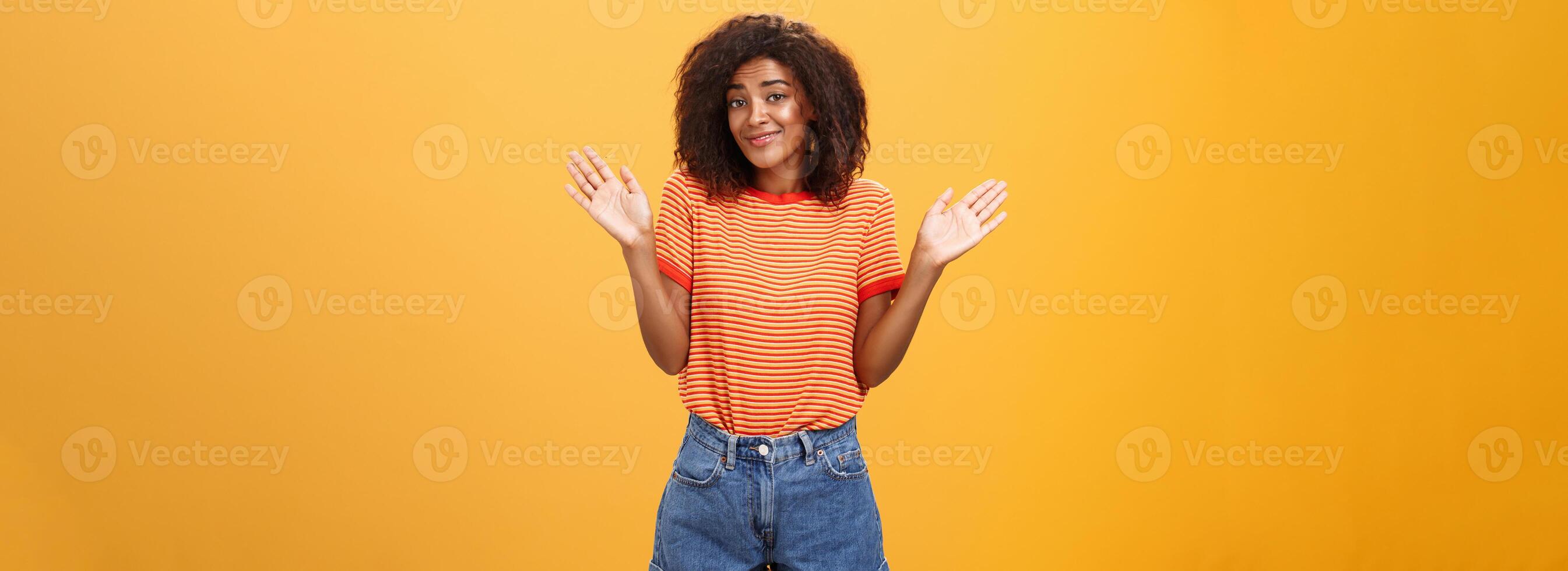 non idée faire ne pas se soucier. négligent et indifférent calme content africain américain femme avec frisé cheveux dans été vêtements élevage paumes et haussant les épaules dans non impliqué pose souriant insouciant étant ignorant photo