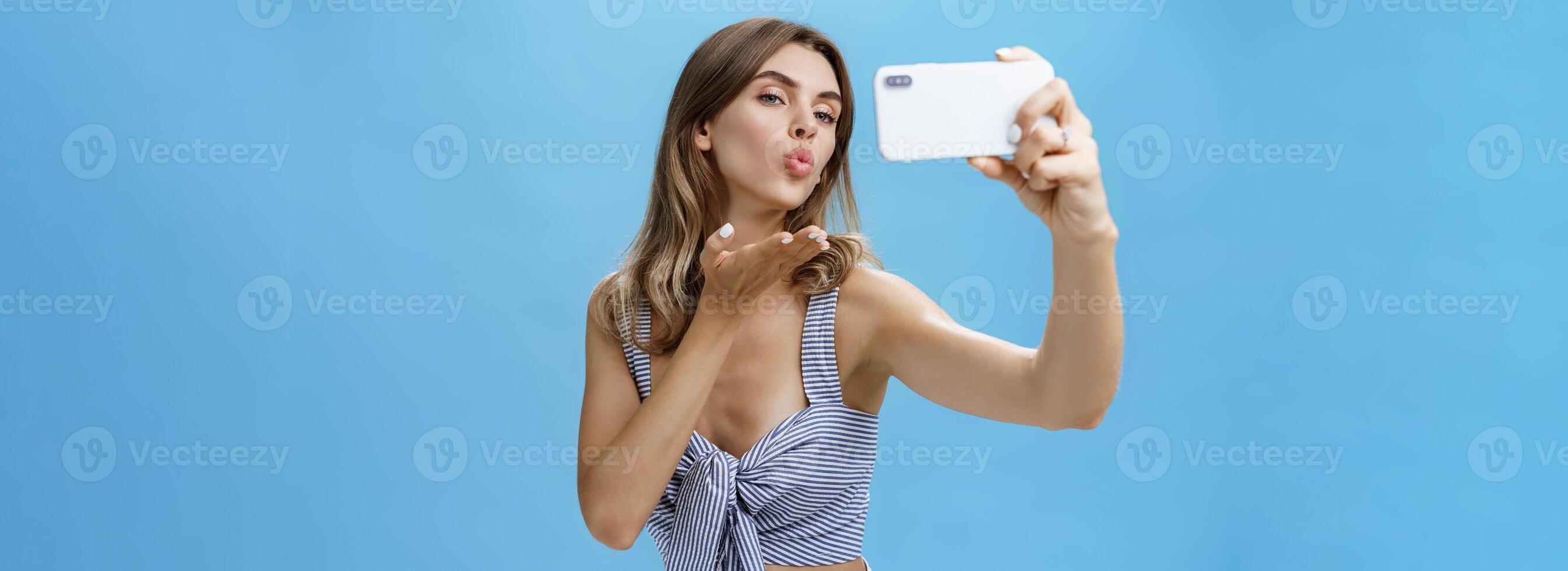 sortant et sûr de lui glamour femme comme prise des photos de se en portant téléphone intelligent fabrication sensuel et affectueux selfie pliant lèvres élevage paume à envoyer vent baiser à écran aimant suiveurs