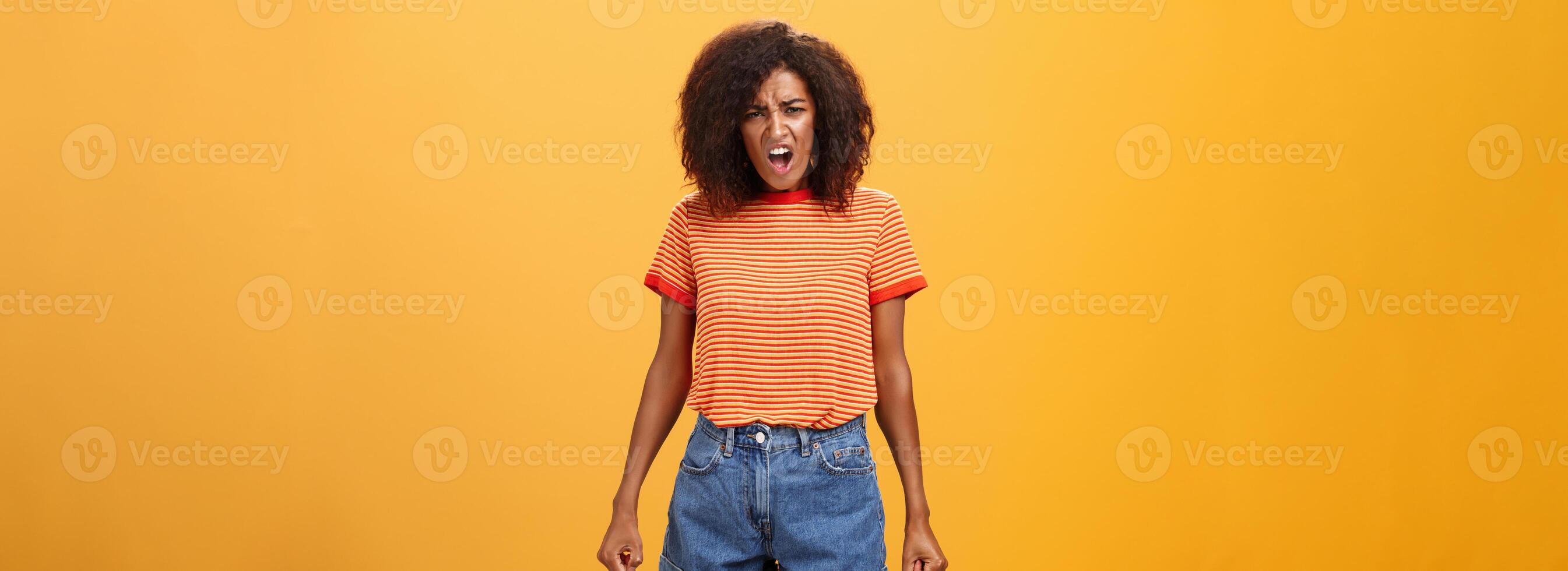 fille se plaindre argumenter avec Parents cause agissant injuste. mécontent énervé et offensé idiot africain américain avec afro coiffure serrant les dents permanent dans insulté pose en hurlant plus de Orange mur photo