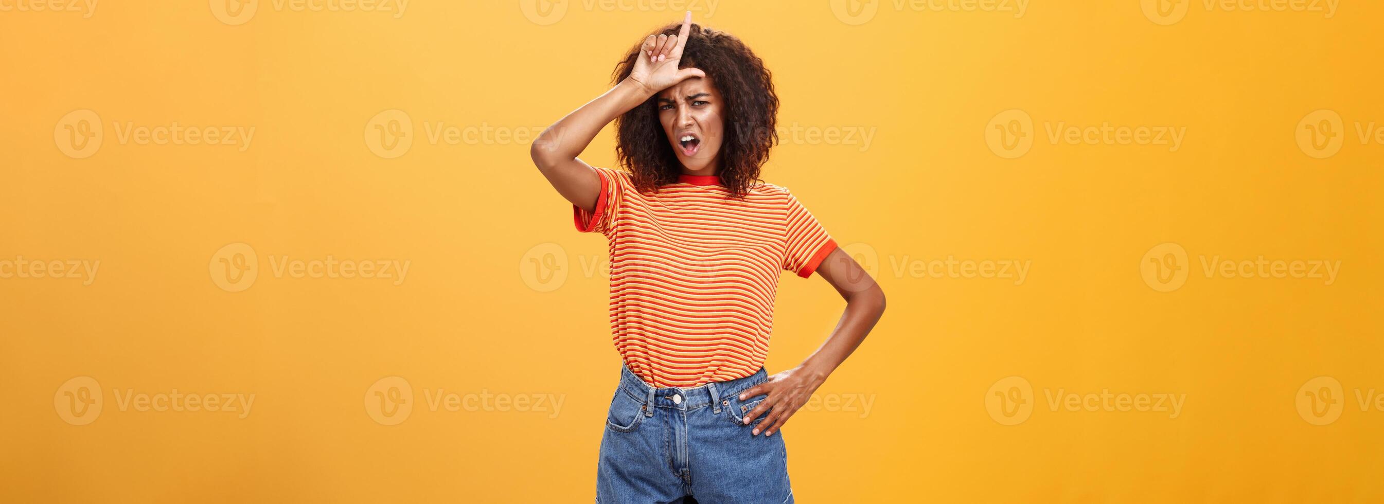 arrogant et sur de soi femme ne pas parler avec perdants. railleur sûr de lui élégant africain américain femelle avec frisé cheveux dans branché vêtements renfrogné réprimande intello, montrant lettre l sur front photo