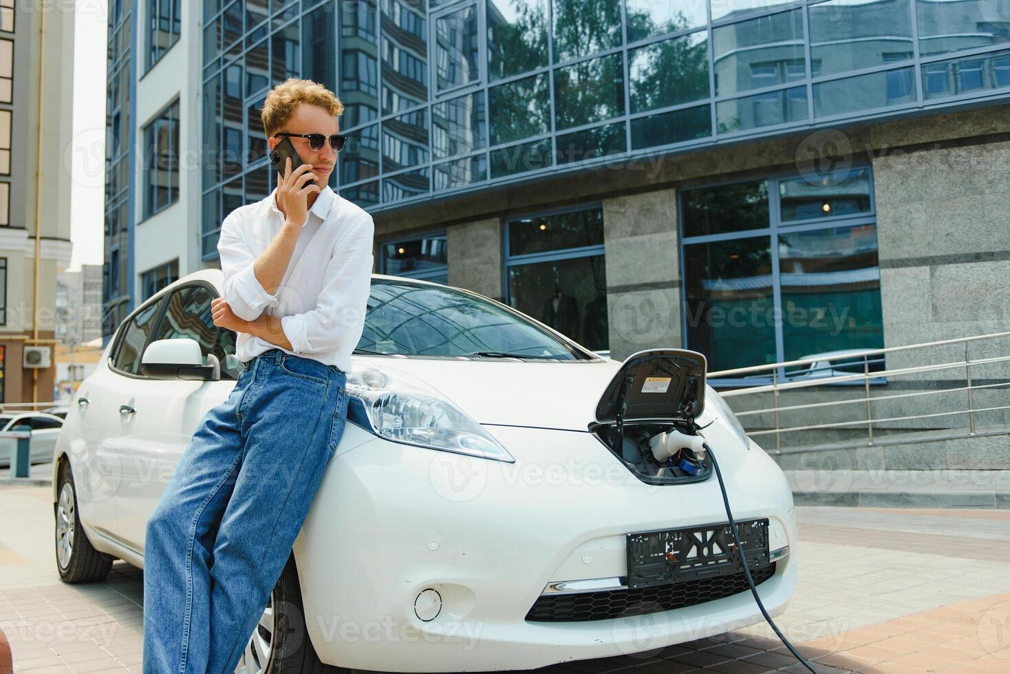 le gars Sam vers le bas sur le capuche de le auto. le sien voiture est mise en charge à le mise en charge gare. une homme regards à le téléphone intelligent écran et sourit. photo