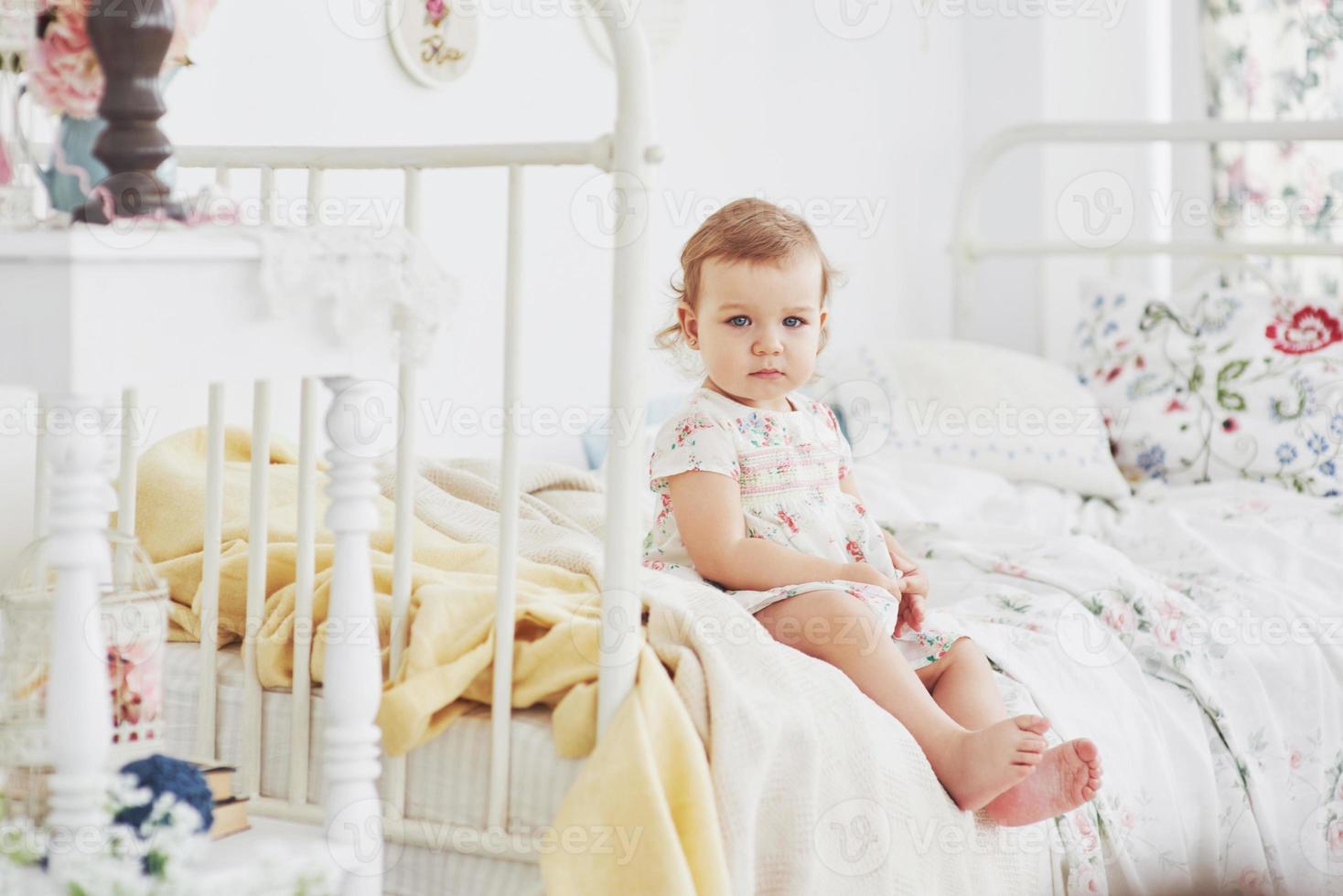 petite fille en robe mignonne implantation au lit jouant avec des jouets à la maison. chambre d'enfant vintage blanche. concept d'enfance photo
