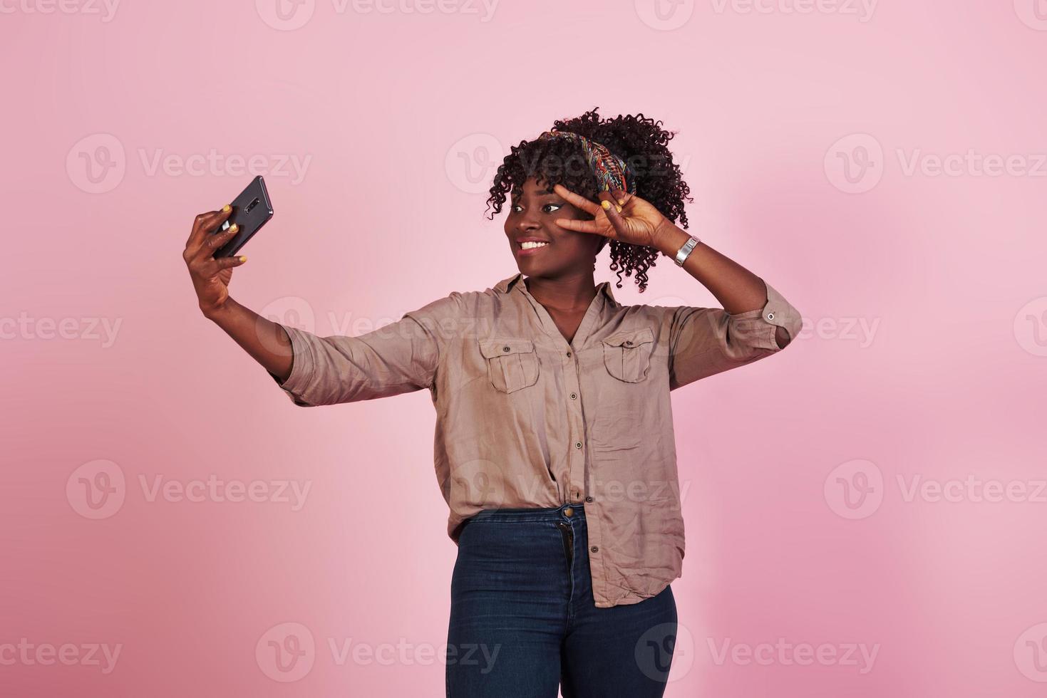 jolie femme afro-américaine en vêtements décontractés prend un selfie et montre un geste avec deux doigts sur fond rose dans le studio photo