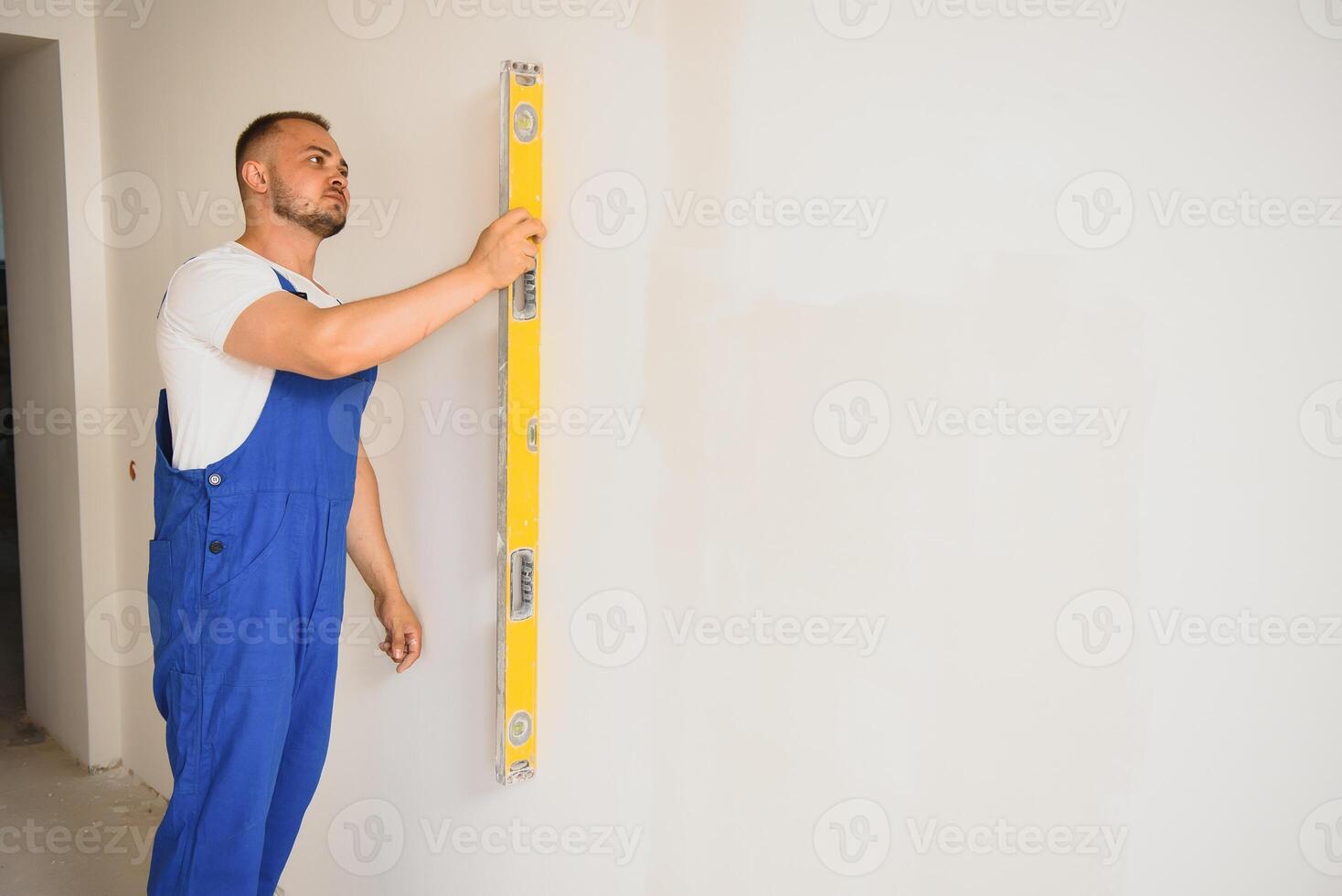 le constructeur soigneusement corrige le irrégularités de le mur avec une truelle. constructeur dans travail vêtements contre une gris mur. photo plâtrier à travail.