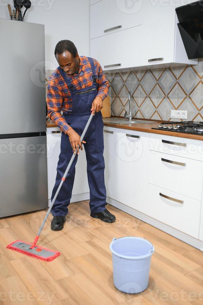 Jeune africain homme lavages le sol avec une balai dans le pièce photo