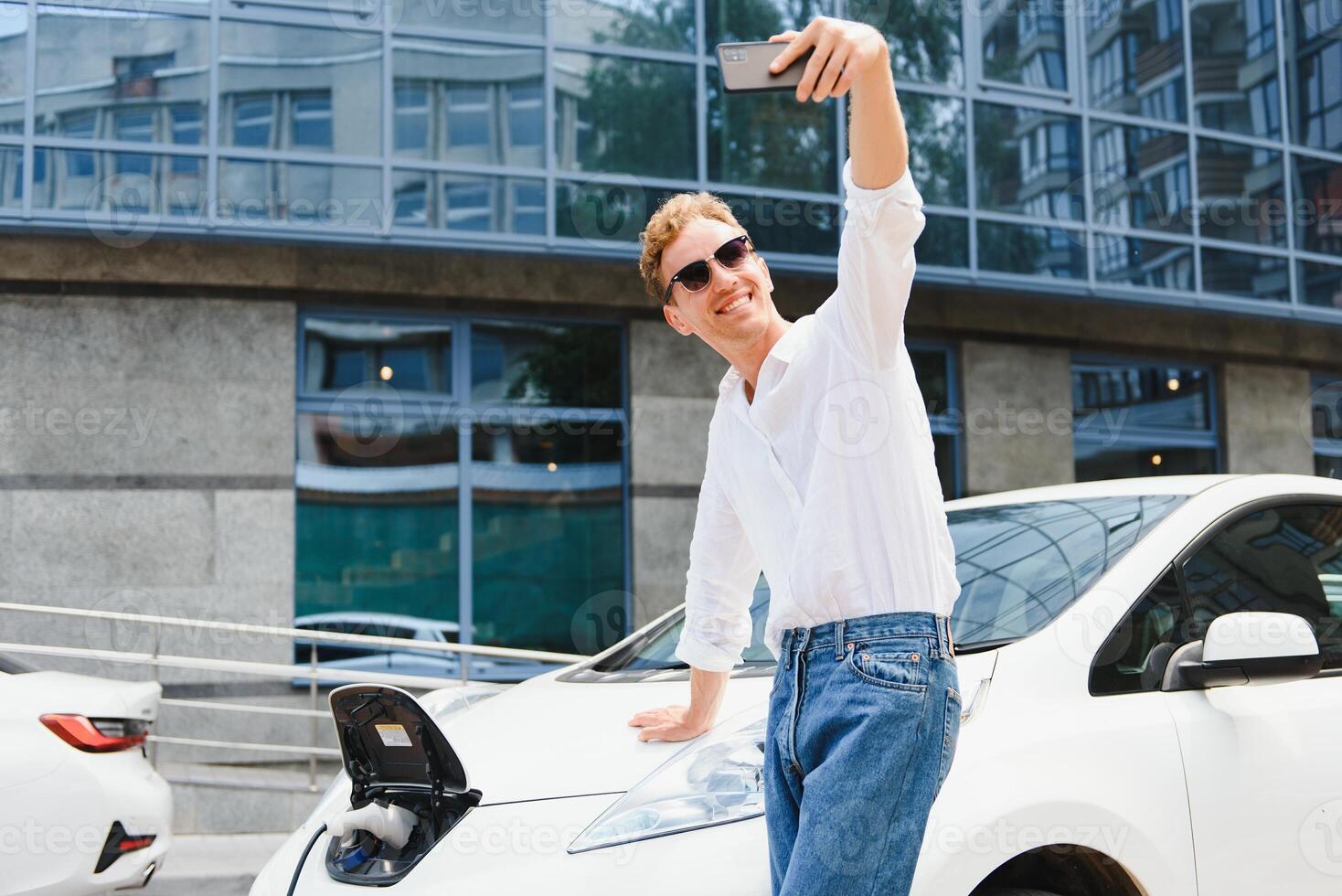 portrait de Jeune Beau européen homme, fabrication selfie photo sur le sien téléphone intelligent tandis que penché sur le sien électrique voiture, mise en charge le batterie à ville Puissance station après achat frais des produits dans Commerce centre commercial