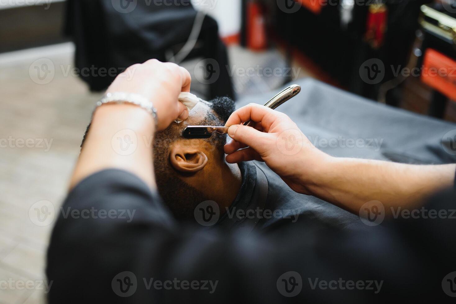 côté vue de sérieux homme avec élégant moderne la Coupe de cheveux à la recherche vers l'avant dans coiffeur magasin. main de coiffeur en gardant tout droit le rasoir et Coupe branché rayures sur tête de client. photo