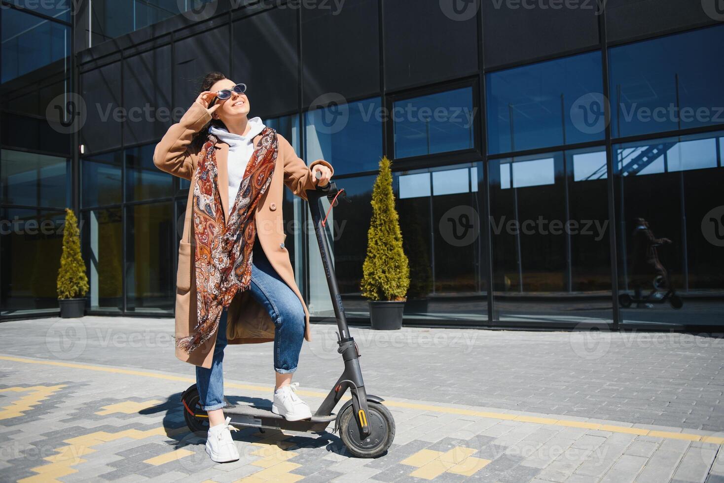 Jeune magnifique femme dans une veste sourit et monte un électrique scooter à travail le long de Bureau bâtiments photo