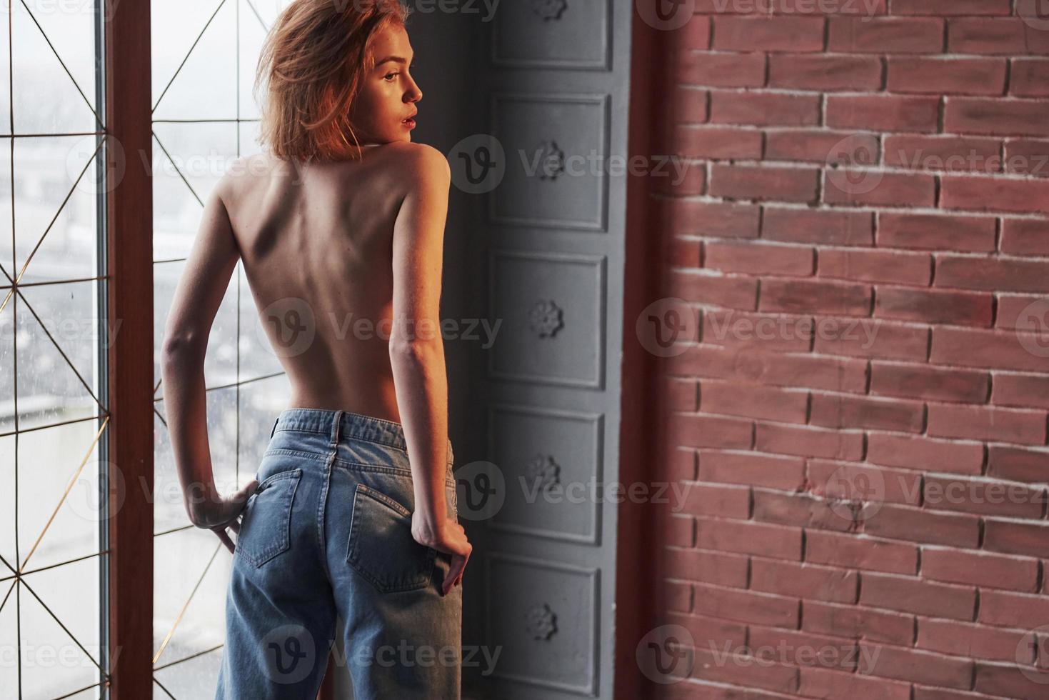 photo de dos. mains sur les poches. jeune blonde chaude avec la poitrine nue et un jean se tient contre la fenêtre