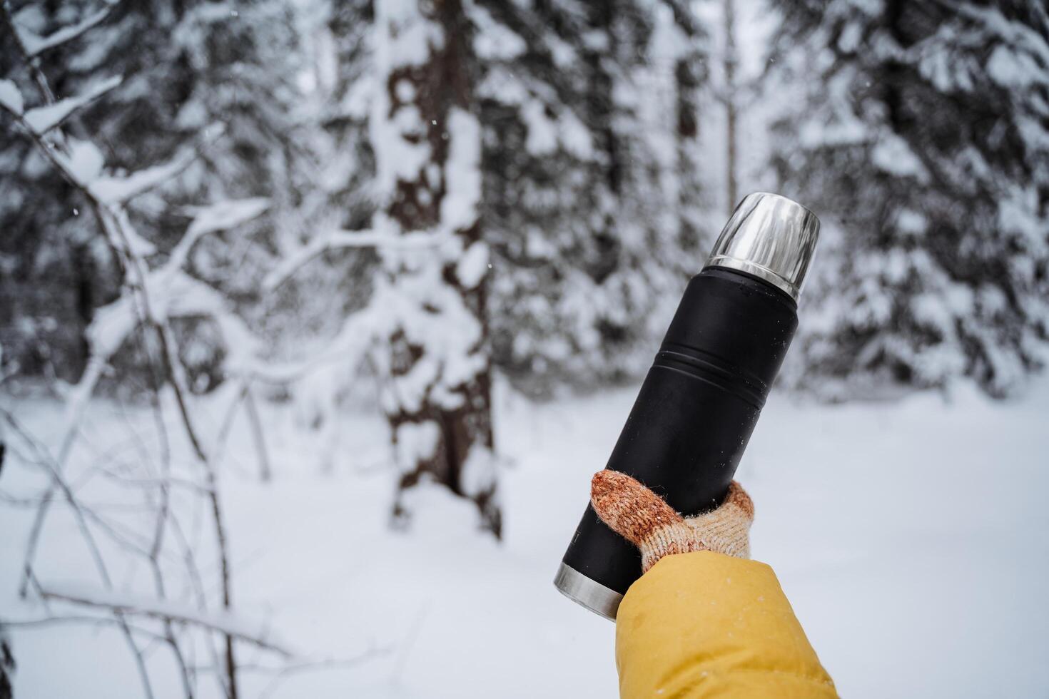 noir mat thermos dans main. voyageur avec gants. boisson chaud thé et café de une thermos sur une marcher dans l'hiver. couvert de neige forêt sur une silencieux journée. brillant vêtements photo