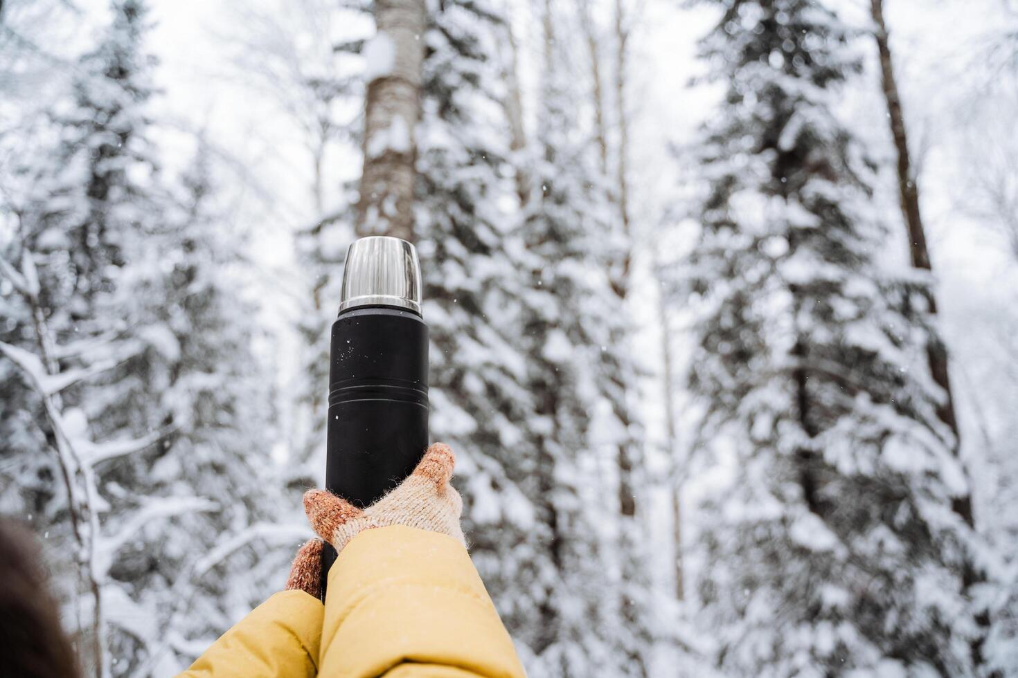noir mat thermos dans main. voyageur avec gants. boisson chaud thé et café de une thermos sur une marcher dans l'hiver. couvert de neige forêt sur une silencieux journée. brillant vêtements photo