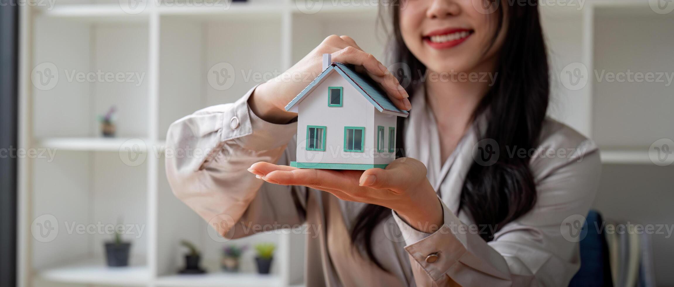 souriant réel biens agent femme mains protégé miniature maison sur en bois bureau. propriété Assurance et honorable la finance calme concept photo