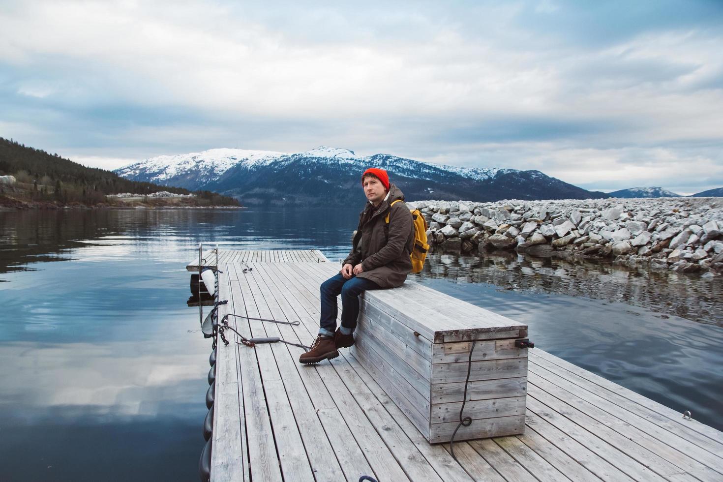 homme voyageur avec un sac à dos jaune portant un chapeau rouge assis sur une jetée en bois sur le fond de la montagne et du lac. espace pour votre message texte ou contenu promotionnel photo