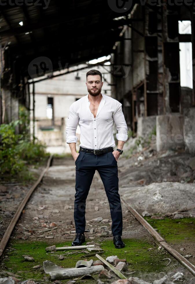 Beau homme est permanent dans déboutonné blanc chemise près cassé béton mur. abandonné entrepôt sur le Contexte photo