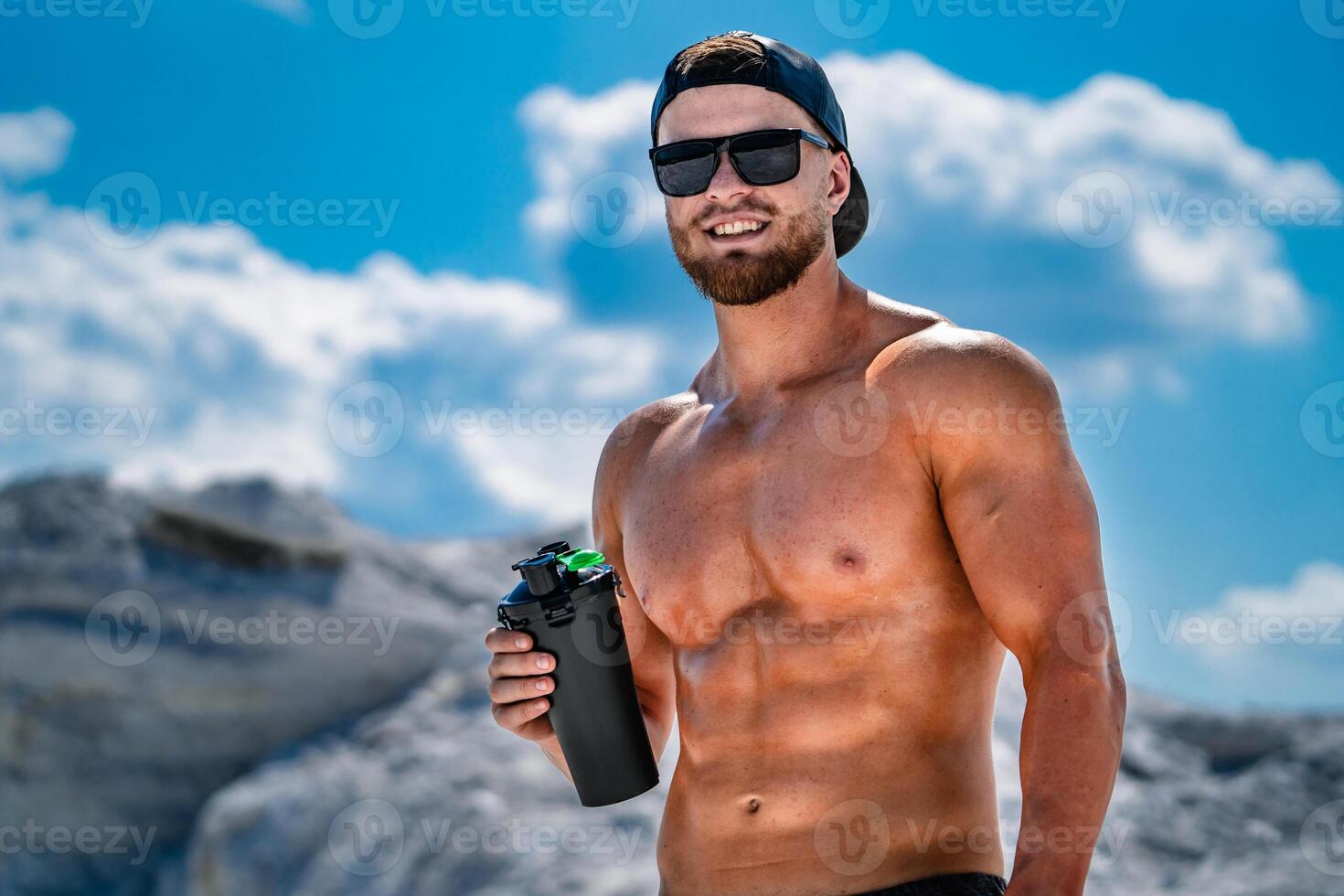 portrait de une Beau à moitié nu bodybuilder homme. des lunettes et une casquette sur. sport bouteille de l'eau dans mains. photo