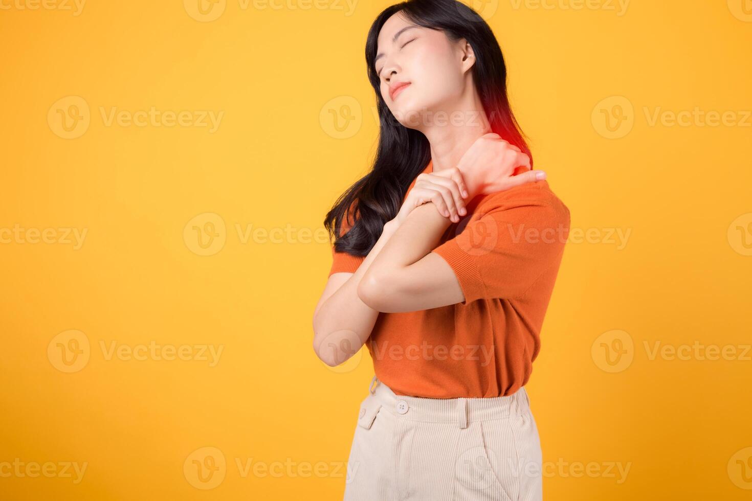 vigilant asiatique femme dans sa 30s, portant un Orange chemise, détient sa douloureux cou sur Jaune Contexte. Bureau syndrome santé se soucier concept. photo