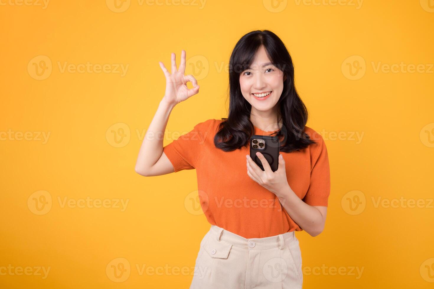 rayonner joie avec une Jeune asiatique femme dans sa 30s, portant un Orange chemise, montrant d'accord main signe tandis que en utilisant téléphone intelligent sur Jaune Contexte. Nouveau app concept. photo