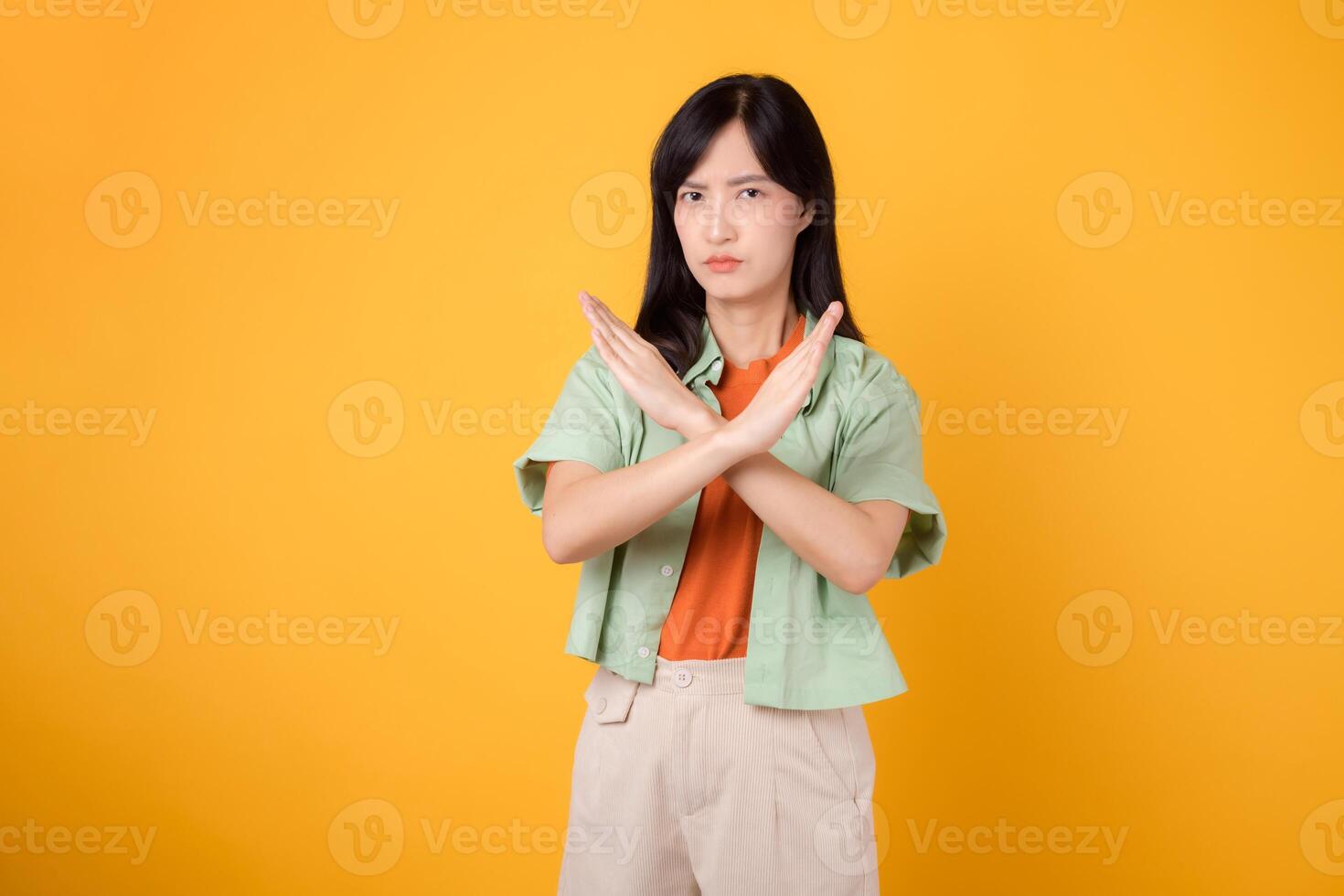 le déni avec Jeune asiatique femme 30s, élégamment vêtu dans un Orange chemise et vert sauteur. sa traverser main geste, ensemble contre une ensoleillé Jaune arrière-plan, transmet le concept de refus et négation. photo