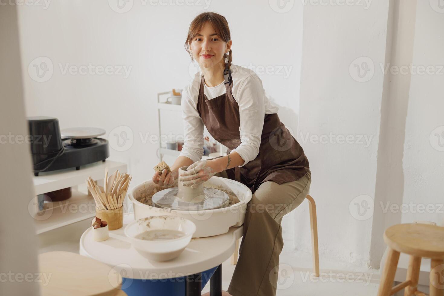 souriant femelle artisan dans tablier séance sur banc avec poterie roue et fabrication argile pot photo