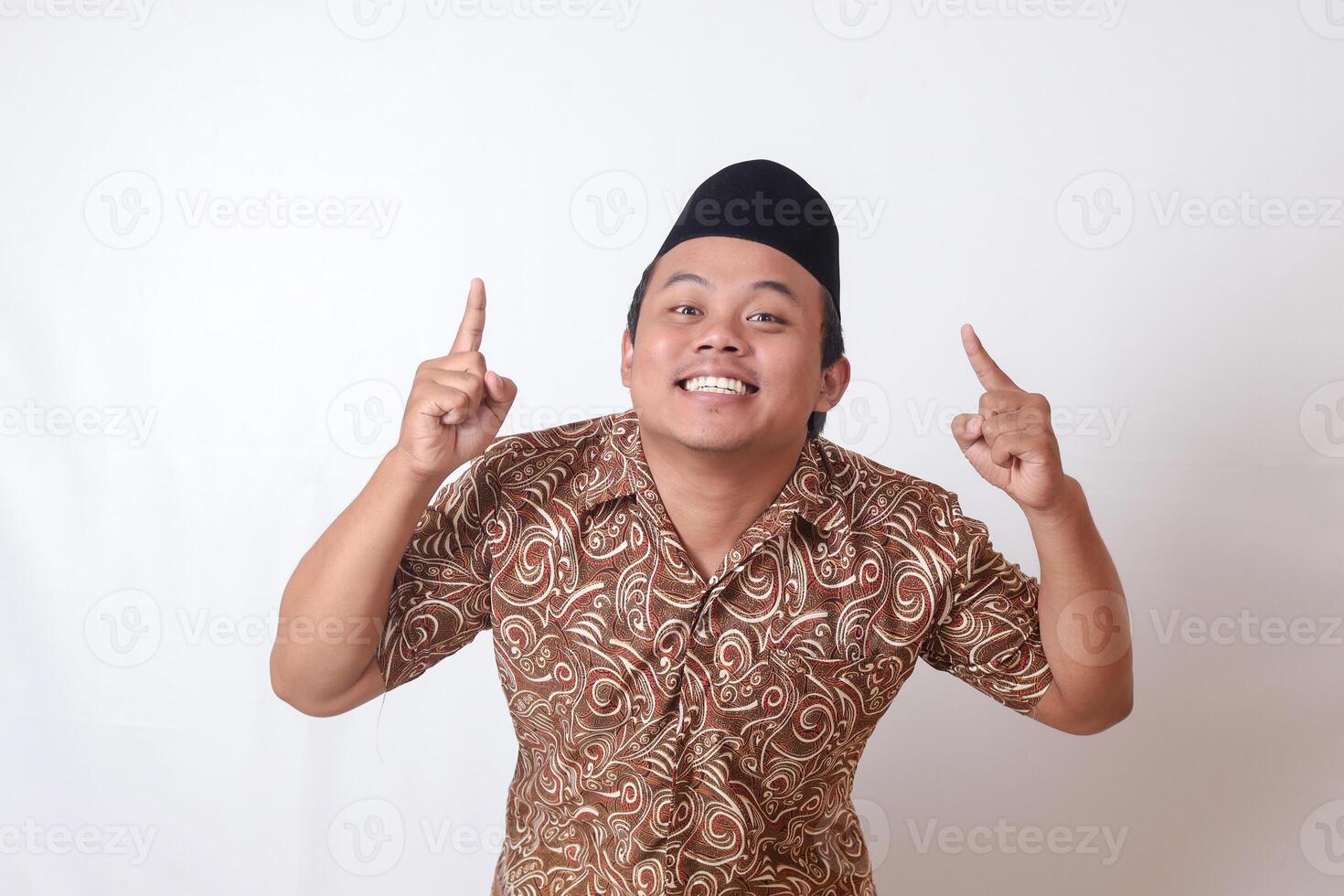 portrait de excité asiatique homme portant batik chemise et Songkok souriant et à la recherche à le caméra montrer du doigt avec deux mains et les doigts à en haut. isolé image sur gris Contexte photo