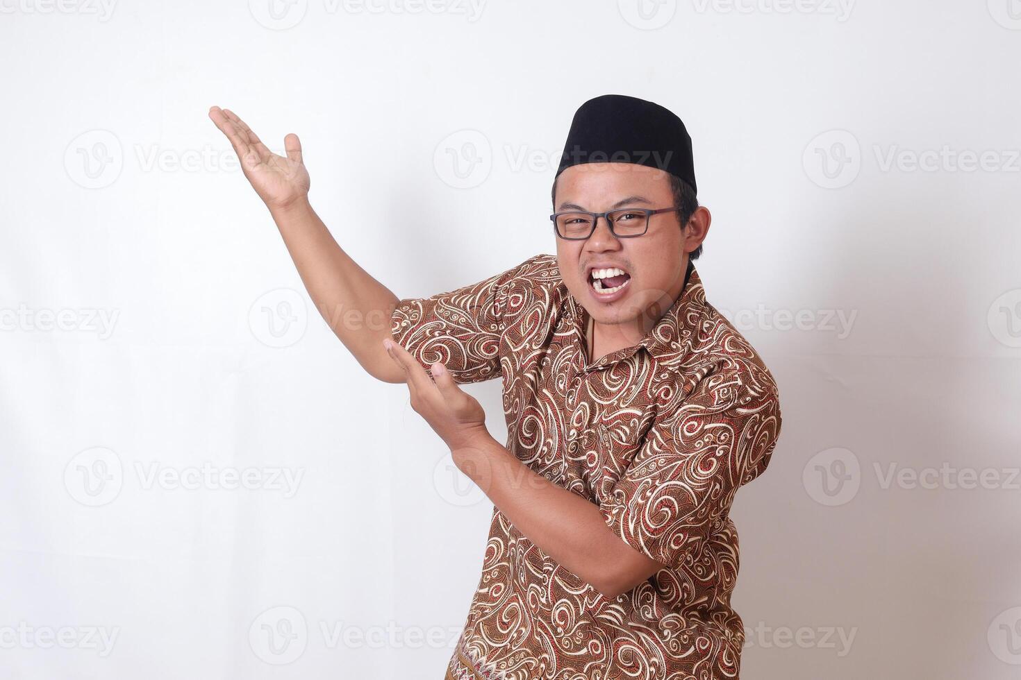 portrait de excité asiatique homme portant batik chemise et Songkok souriant et à la recherche à le caméra montrer du doigt avec deux mains et les doigts à le côté. isolé image sur gris Contexte photo