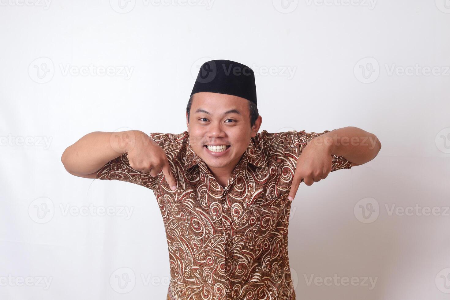 portrait de excité asiatique homme portant batik chemise et Songkok souriant et à la recherche à le caméra montrer du doigt avec deux mains et les doigts à le vers le bas côté. isolé image sur gris Contexte photo