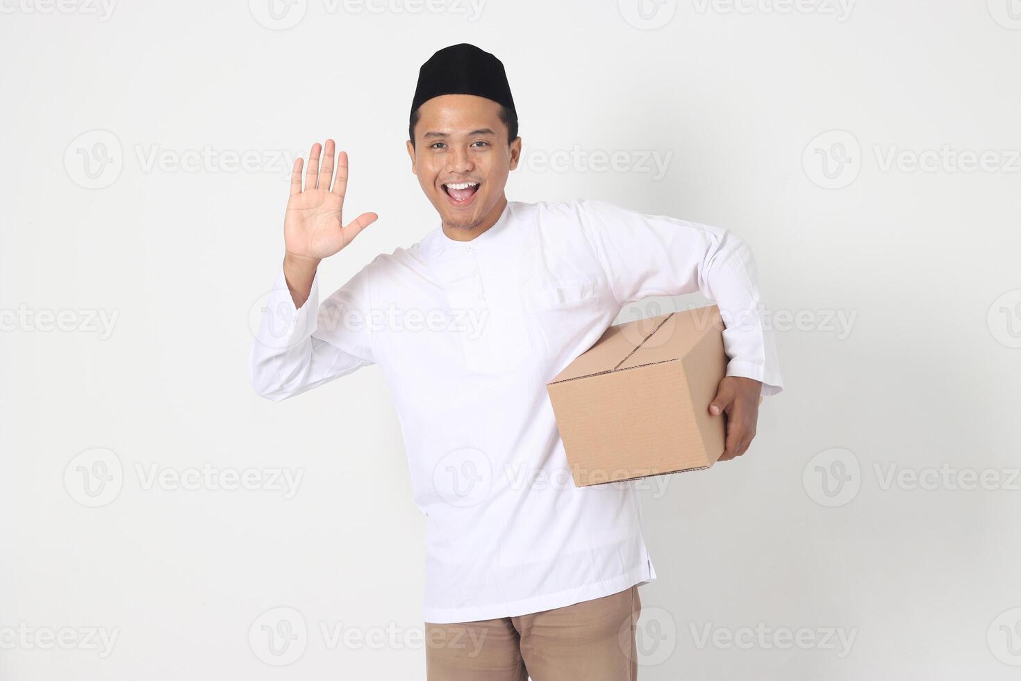 portrait de excité asiatique musulman homme dans koko chemise avec peci en disant Salut, salutation à Quelqu'un tandis que porter papier carton boîte. Aller Accueil pour eid moubarak. isolé image sur blanc Contexte photo