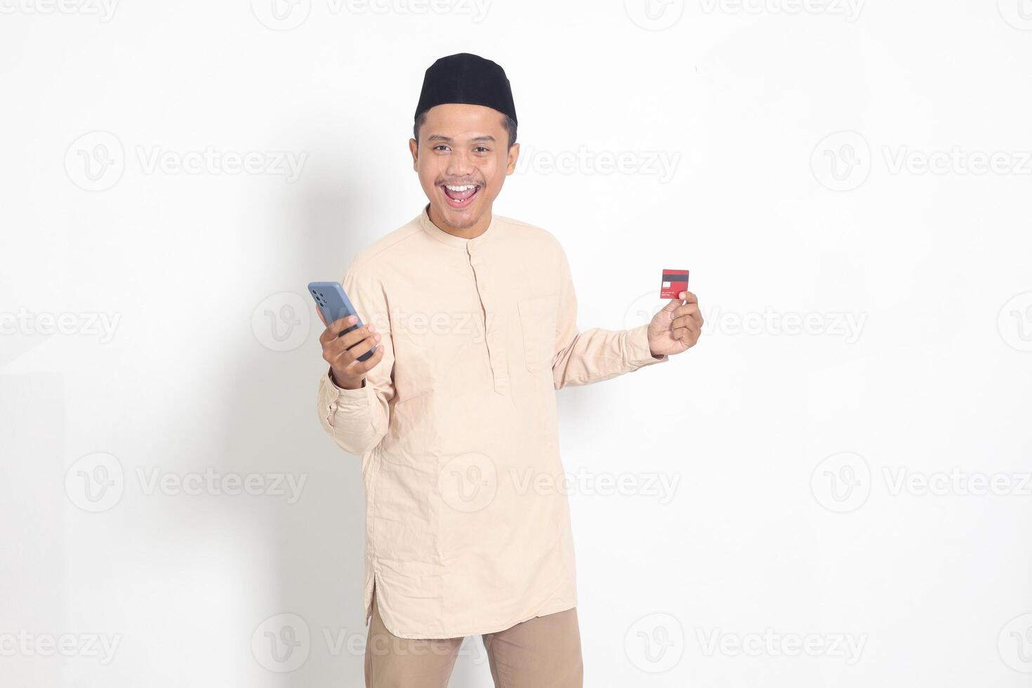 portrait de attrayant asiatique musulman homme dans koko chemise avec calotte en portant une mobile téléphone et en présentant crédit carte. isolé image sur blanc Contexte photo