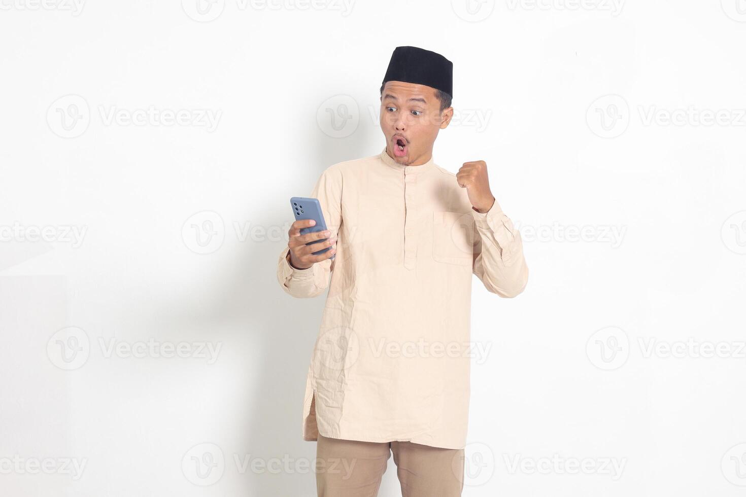 portrait de excité asiatique musulman homme dans koko chemise avec calotte élevage le sien poing, célébrer gagnant Jeu sur mobile téléphone. La publicité et social médias concept. isolé image sur blanc Contexte photo
