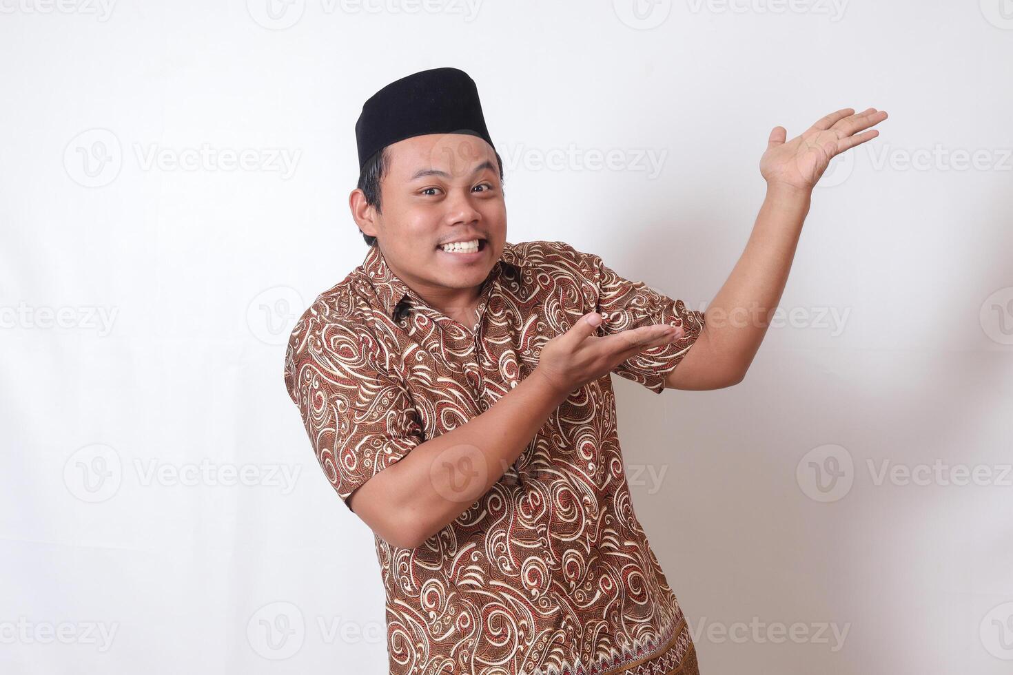 portrait de excité asiatique homme portant batik chemise et Songkok souriant et à la recherche à le caméra montrer du doigt avec deux mains et les doigts à le côté. isolé image sur gris Contexte photo