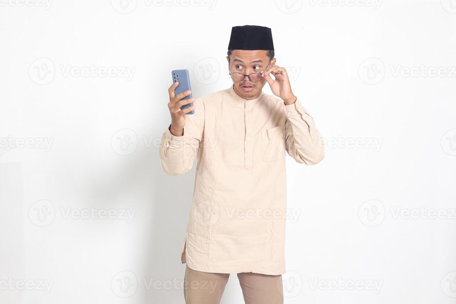 portrait de surpris asiatique musulman homme dans koko chemise avec calotte en portant mobile téléphone, montrant sensationnel sous le choc visage expression. La publicité et social médias concept. isolé image sur blanc Contexte photo