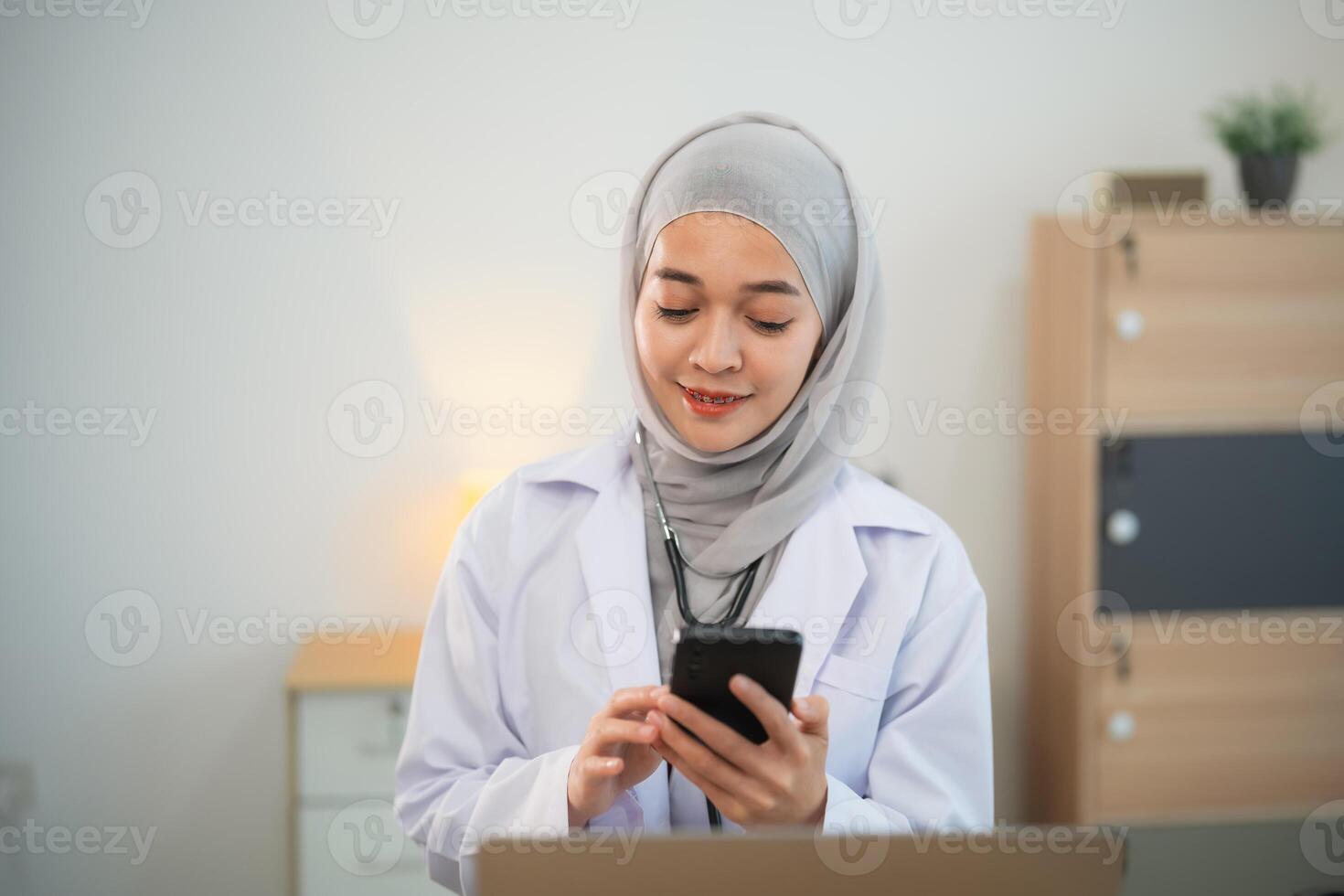 musulman Islam asiatique médecin homme portant hijab et stéthoscope travail avec portable ordinateur et en utilisant mobile téléphone, téléphone intelligent. femelle médecin travail à Accueil Bureau hôpital. santé hôpital concept. photo