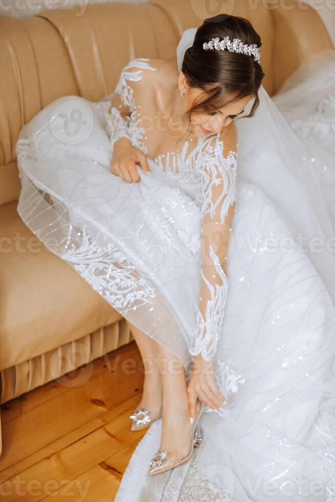 Jeune la mariée dans magnifique mariage robe en mettant sur des chaussures à l'intérieur. la mariée Robes des chaussures avant le mariage cérémonie. détail de la mariée en mettant sur haute talon sandale mariage chaussures. mariage la mariée chaussures. photo