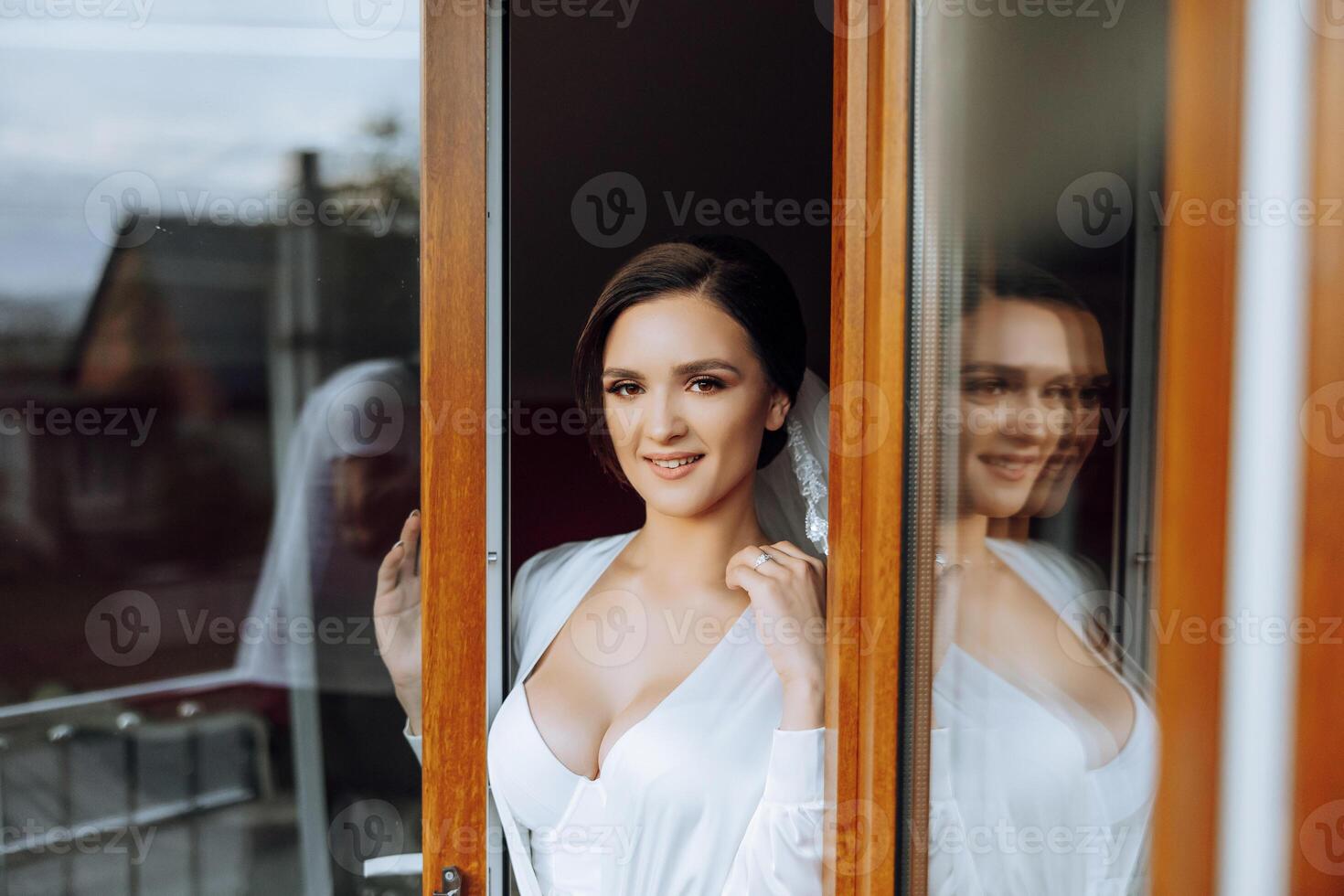 le la mariée des stands sur le balcon entre le ouvert les fenêtres dans une blanc peignoir et regards devant. mariage journée. une magnifique la mariée. mariage. photo