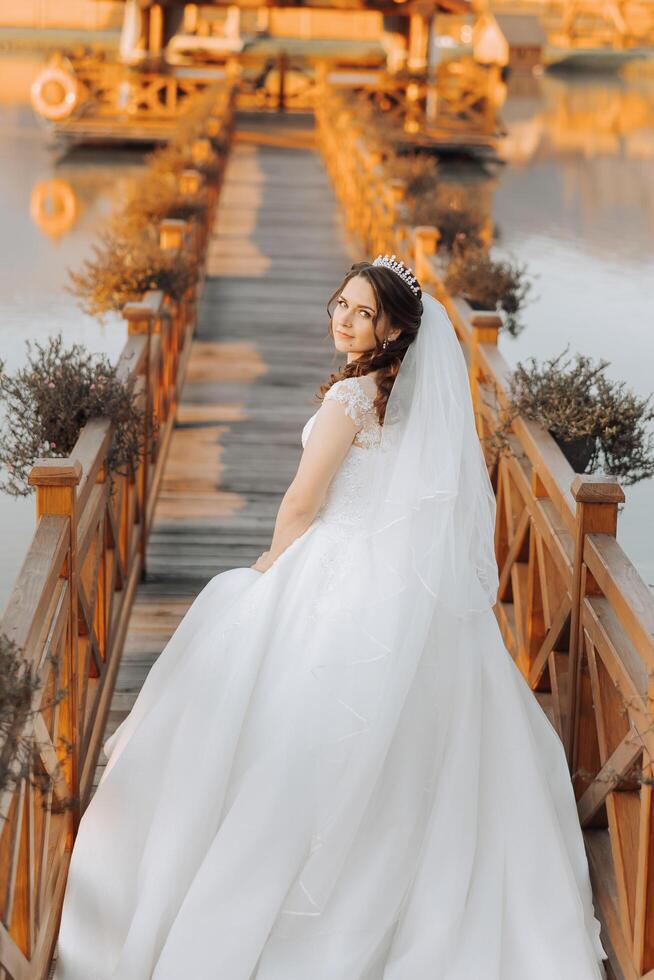 une brunette la mariée dans une blanc robe avec une longue train détient le robe et des promenades sur une en bois pont. l'automne. mariage photo session dans la nature. magnifique cheveux et se maquiller. fête